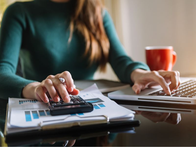 A professional woman in a green sweater working on financial documents with a calculator and a laptop on a desk. A red coffee mug sits beside her, indicating a work-focused environment.