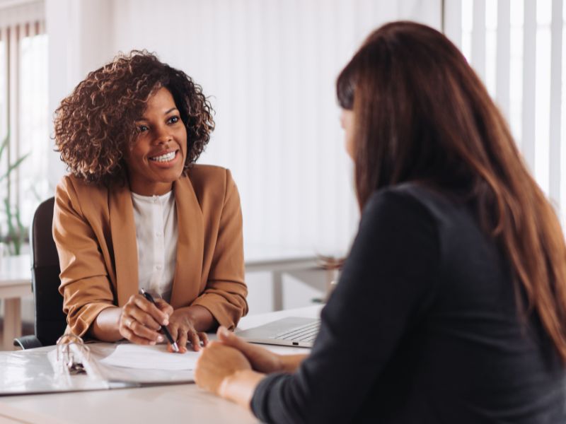 A wider view of a financial consultation session where a consultant and client are engaged in reviewing paperwork and discussing financial reports. A laptop is visible in the background, enhancing the professional setting.