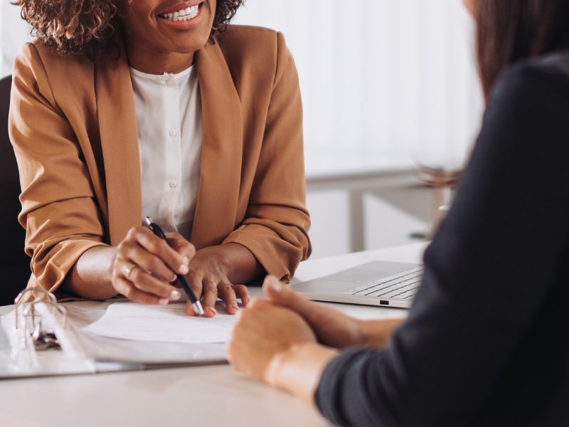 A financial consultant in a brown blazer engaging in a discussion with a client. She is smiling while holding a pen and reviewing documents, suggesting a professional and collaborative consultation.