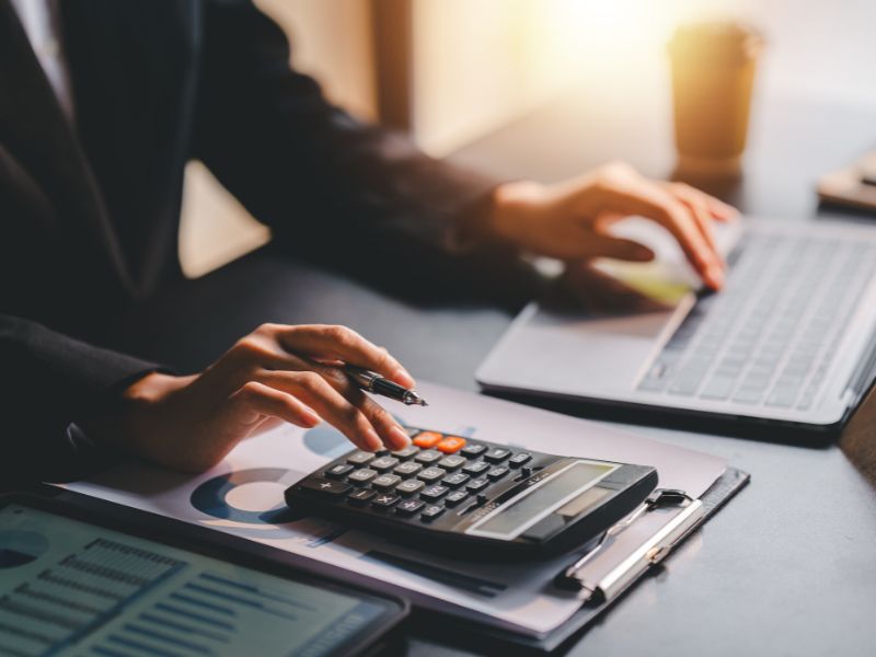 A close-up of hands typing on a laptop while using a calculator to manage bookkeeping tasks. Financial reports with graphs and charts are spread out on the table.