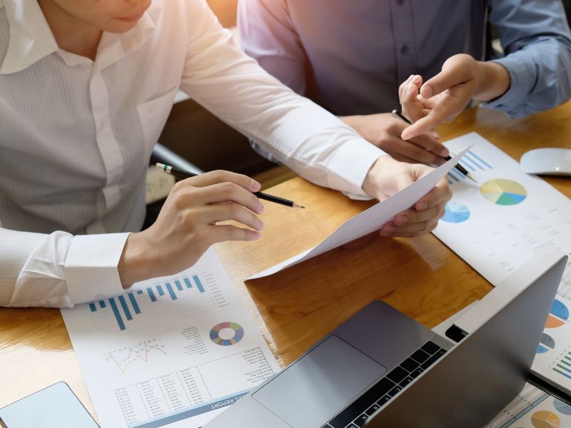 Two business professionals analyzing a financial report together. One person points at a document with a pen while another takes notes, showing teamwork in financial planning.
