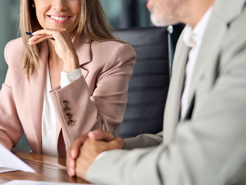 A senior financial consultant, dressed in a beige suit, is engaged in a serious financial discussion with a client. She holds a financial report and is poised to provide strategic insights. The client, dressed in professional business attire, listens attentively while resting his hands on the table, indicating engagement in the conversation. The setting, with its modern office furniture and clean workspace, underscores the high level of expertise and trust involved in CFO advisory services, financial planning, and corporate decision-making.