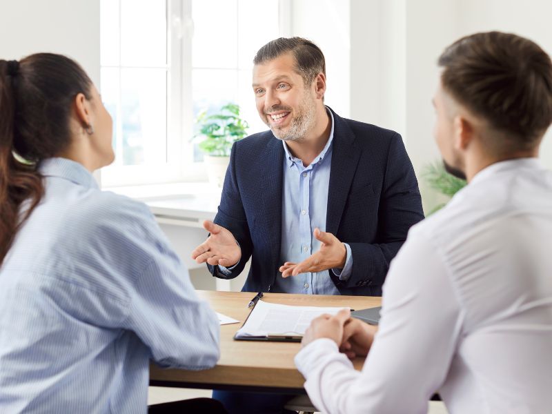 A smiling business consultant dressed in a beige blazer is engaged in a financial discussion with a client at a modern office. She holds a document in one hand and a pen in the other, appearing confident and professional. The well-lit workspace suggests an environment of trust and expertise. The conversation appears to center around financial planning, tax strategies, or investment consulting, highlighting the role of an experienced CFO in guiding businesses through complex financial landscapes.