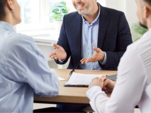 A close photo of a smiling business consultant dressed in a beige blazer is engaged in a financial discussion with a client at a modern office. She holds a document in one hand and a pen in the other, appearing confident and professional. The well-lit workspace suggests an environment of trust and expertise. The conversation appears to center around financial planning, tax strategies, or investment consulting, highlighting the role of an experienced CFO in guiding businesses through complex financial landscapes.
