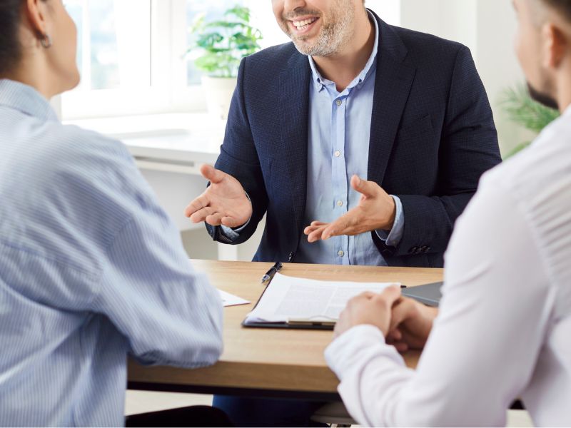 LedgerLogix Experienced CFO Services Close A close photo of a smiling business consultant dressed in a beige blazer is engaged in a financial discussion with a client at a modern office. She holds a document in one hand and a pen in the other, appearing confident and professional. The well-lit workspace suggests an environment of trust and expertise. The conversation appears to center around financial planning, tax strategies, or investment consulting, highlighting the role of an experienced CFO in guiding businesses through complex financial landscapes.