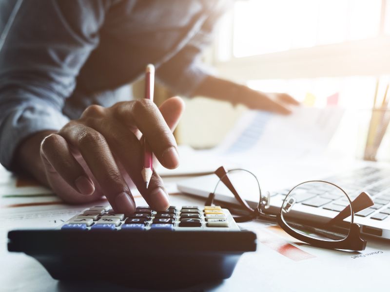A financial professional is seated at a desk, carefully using a calculator while cross-referencing numbers on printed financial documents. The workspace is filled with spreadsheets, data charts, and graphs, emphasizing the importance of precise financial calculations. A pair of reading glasses rests on the table, signifying a detailed and methodical approach to bookkeeping and accounting. The soft lighting in the image creates a focused atmosphere, reinforcing the theme of financial accuracy and thorough analysis.