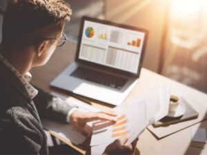 A professional working at a desk is analyzing printed financial reports while viewing profit and revenue trends on a laptop screen. The scene is bathed in warm sunlight, symbolizing strategic decision-making and financial optimization through data-driven insights.