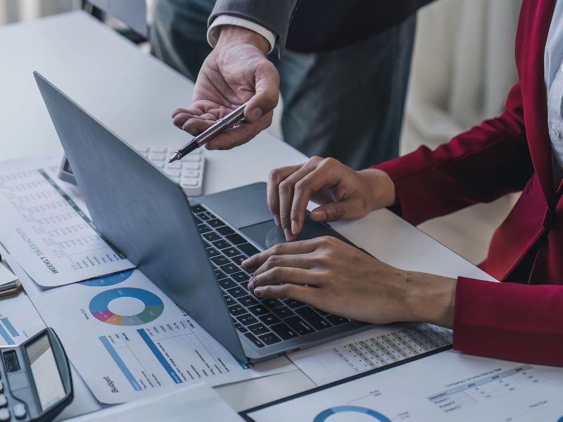 A collaborative business meeting where one professional is pointing at a laptop displaying financial performance graphs while another takes notes. The discussion revolves around reviewing profitability, making strategic adjustments, and ensuring sustainable business growth.