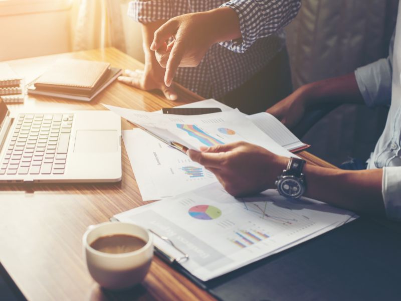 A close-up of hands working on an accounting spreadsheet, with one person using a calculator while another holds a pen. Various financial charts and reports fill the desk, representing financial data analysis, budgeting, and bookkeeping.