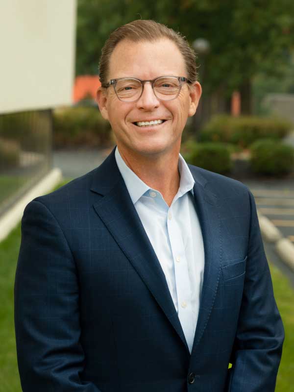 A photo of Todd Pfister, a Partner of TP4 Advisors standing outside the office building wearing a light blue dress shirt with a dark suit.