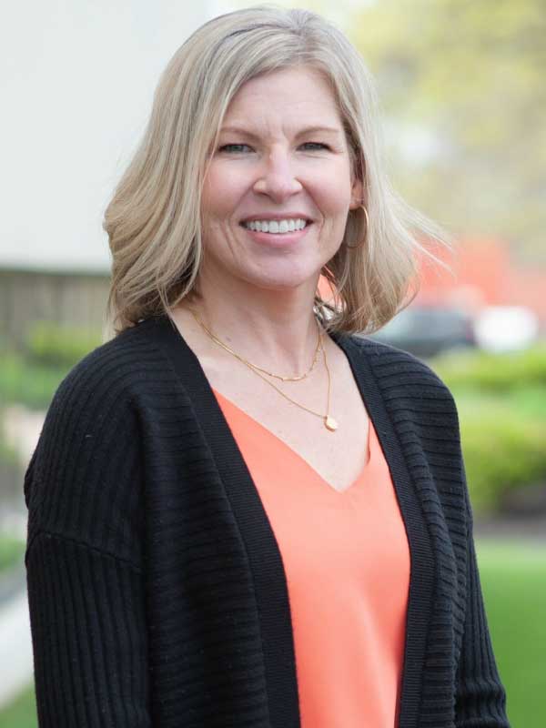 A photo of Tricia Moorman wearing a peach blouse with a black blazer jacket outside TP4 Advisors office building.